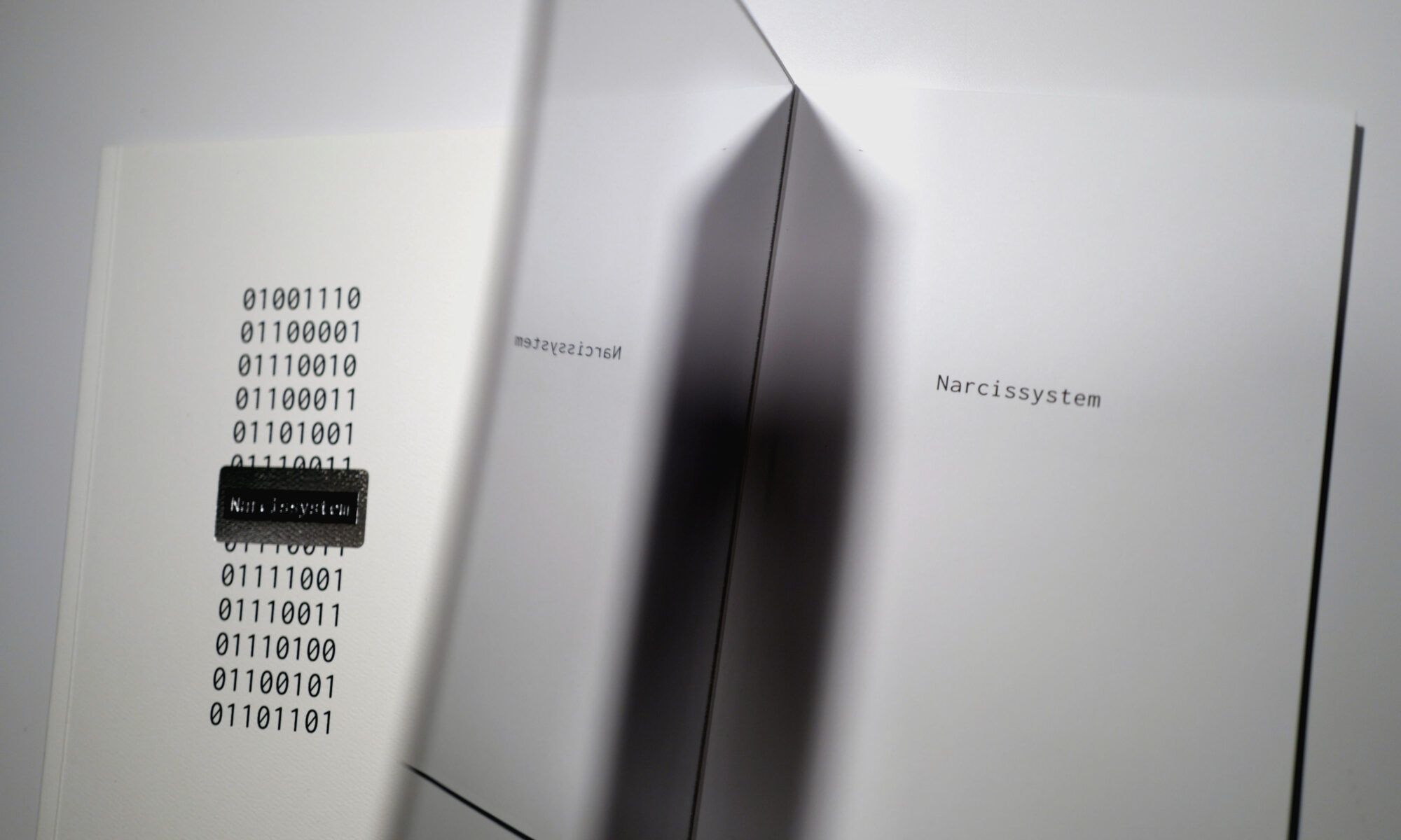 Two copies of Narcissystem, the one on the right opened to show a mirrored inside front cover reflecting the half-title page.